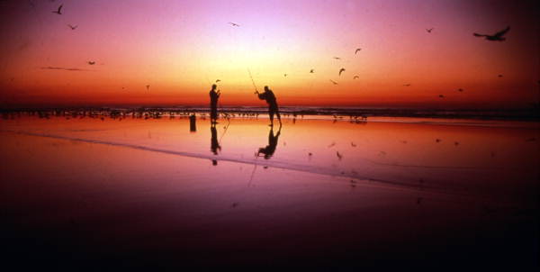 Pêcheurs et oiseaux au lever du soleil sur une grande plage.
