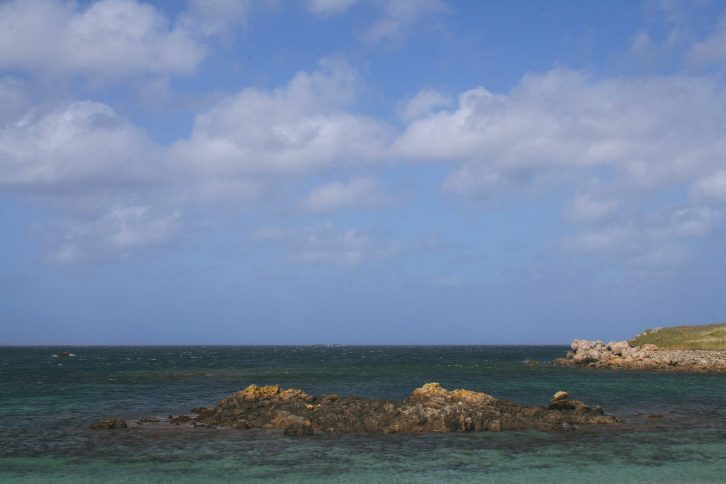 Rocher à marée haute et mer calme sous un ciel bleu parsemé de nuages.