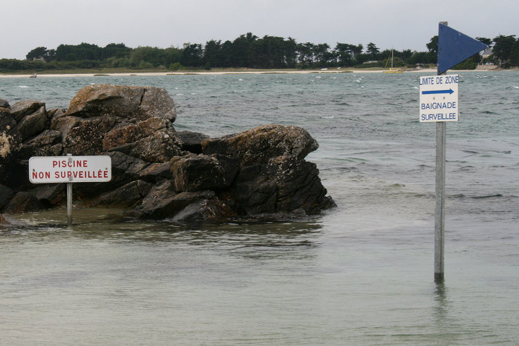 Deux écriteaux disposés dans l'eau au bord d'une plage indiquent respectivement que la piscine n'est pas surveillée, mais que la baignade l'est.
