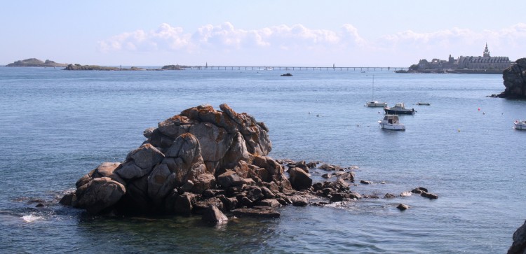 Paysage marin avec rochers, bateau de plaisance et, au loin, la ville de Roscoff et la passerelle de l'estacade qui permet, à marée basse, d'embarquer pour l'île de Batz.