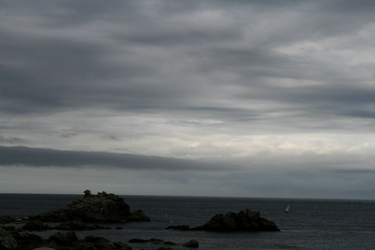 Ciel chargé de nuages gris sur une mer parsemée de gros rochers et parcourue par un petit voilier.