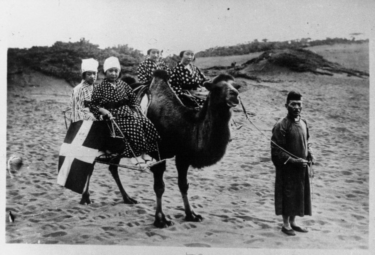 Dans le désert, un chameau conduit par un homme porte quatre jeunes femmes en costume traditionnel de Mandchourie.