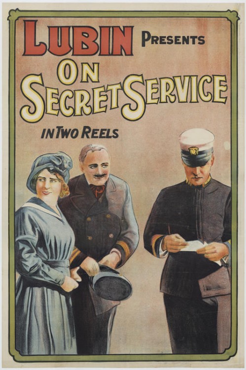 Affiche de film représentant deux hommes et une femme. Les hommes sont en uniforme, la femme porte une robe et un chapeau bleu. L'un des deux hommes est en train de lire ce qui est écrit sur un papier qu'il tient entre ses mains pendant que l'autre le regarde, un sourire énigmatique sur ses lèvres soulignées par une fine moustache.
